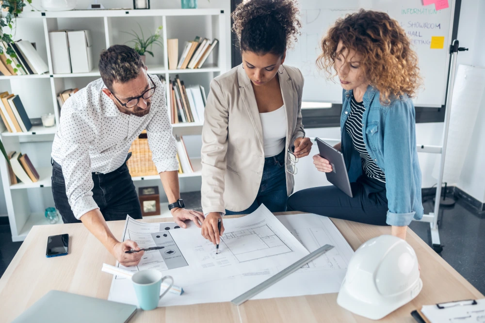 Three colleagues discussing architectural floor plans spread across a wooden office desk.