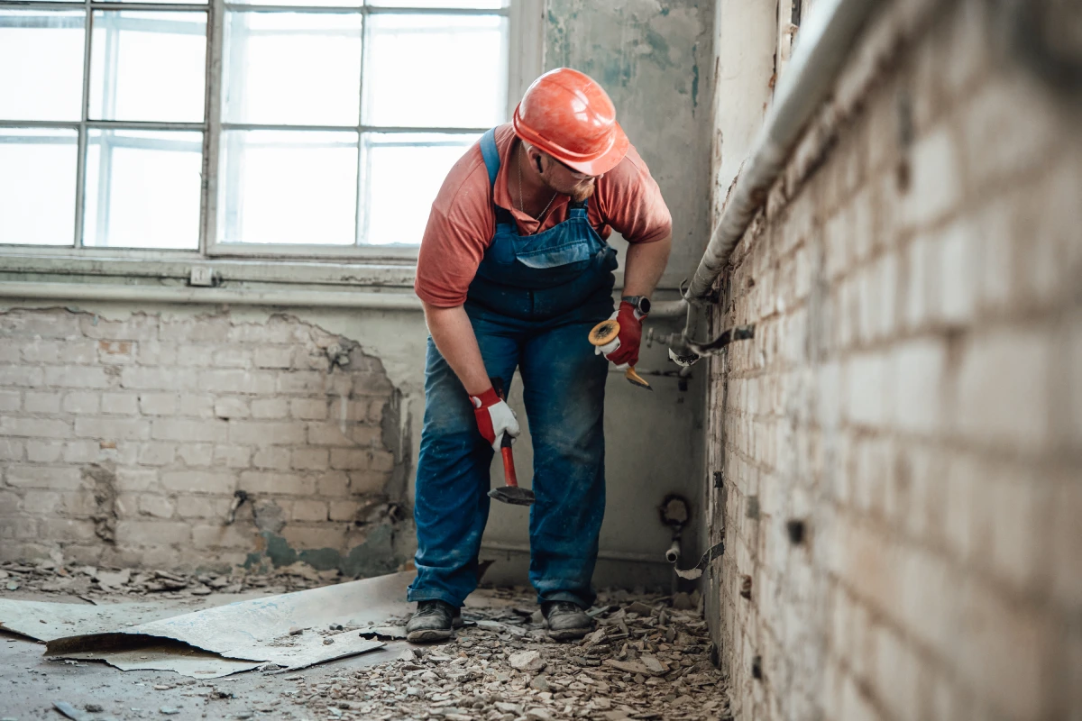 A construction worker wearing an orange hard hat and blue overalls uses a hammer.
