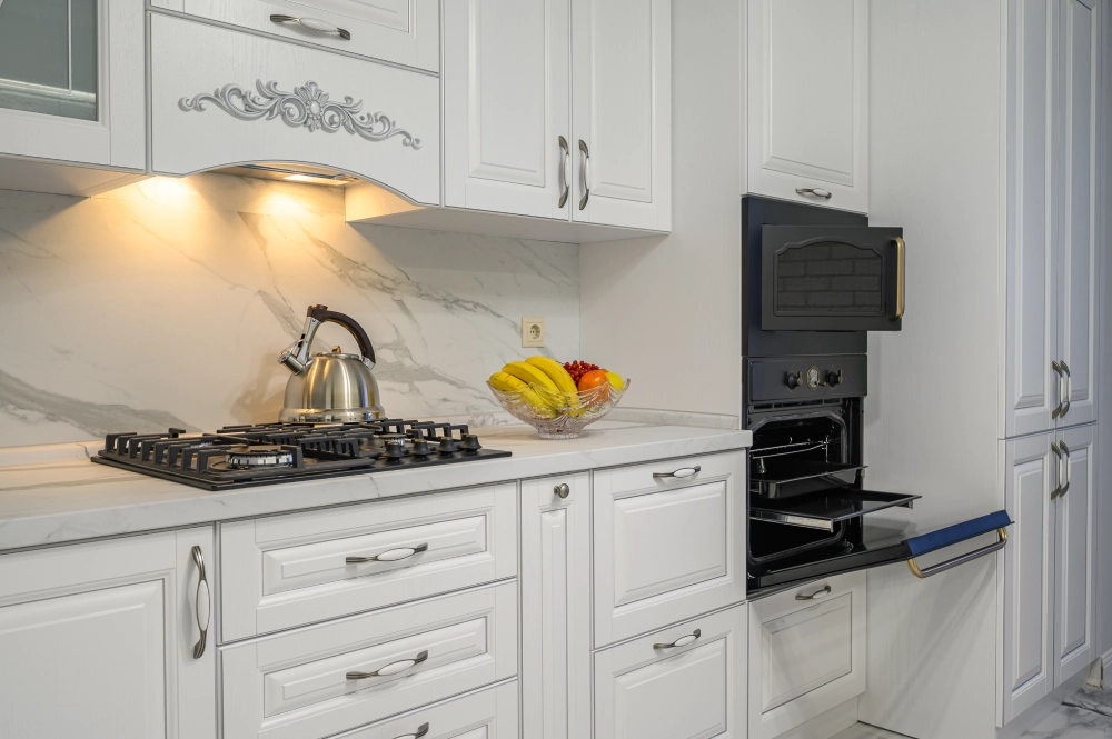 A modern white kitchen featuring a marble backsplash and built-in black oven with open door.