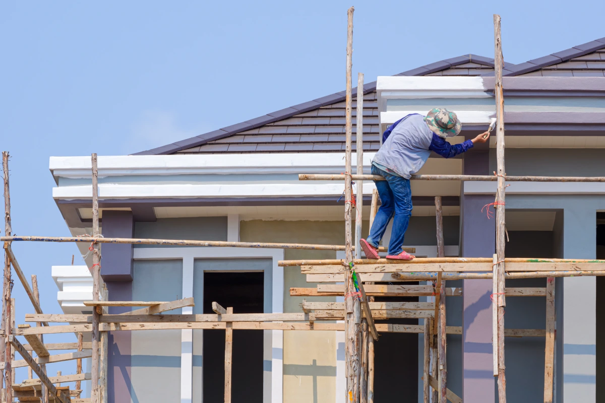 A construction worker on a wooden scaffold paints the trim of a house.
